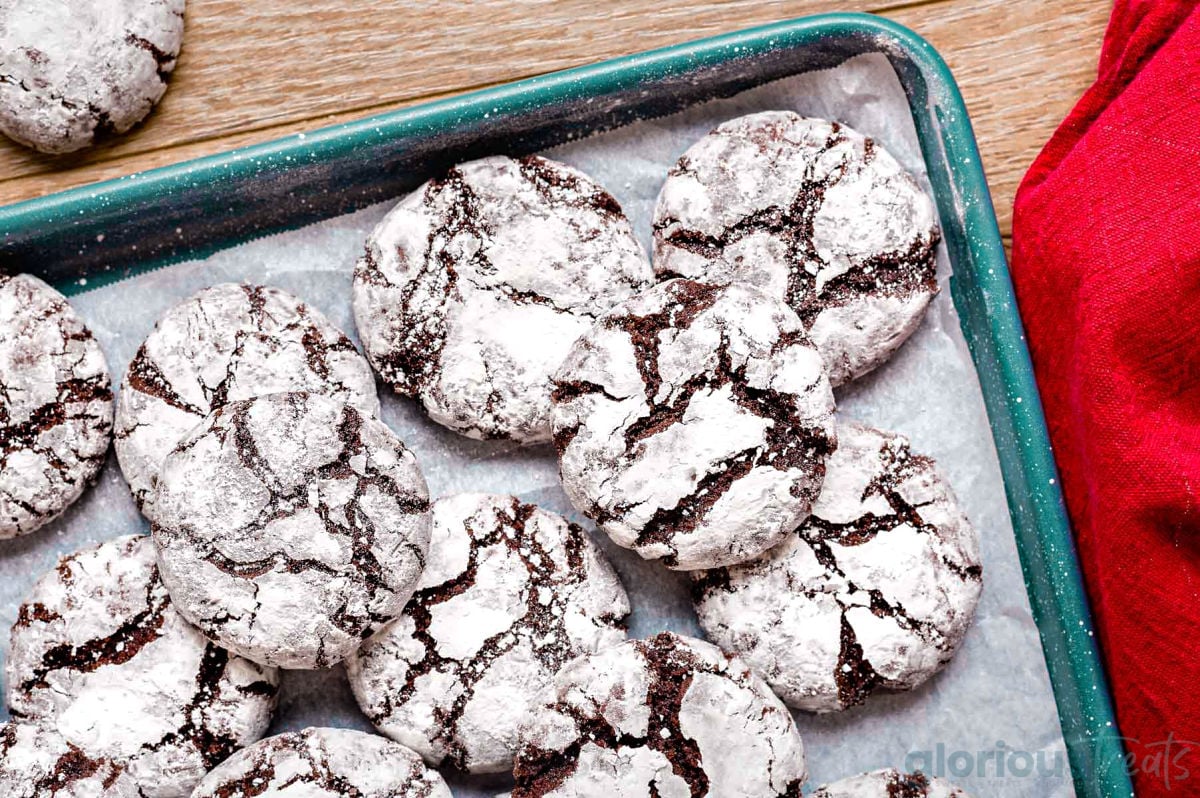 blue baking sheet with a batch of chocolate crinkle cookies on parchment paper on top. sitting next to a red napkin.