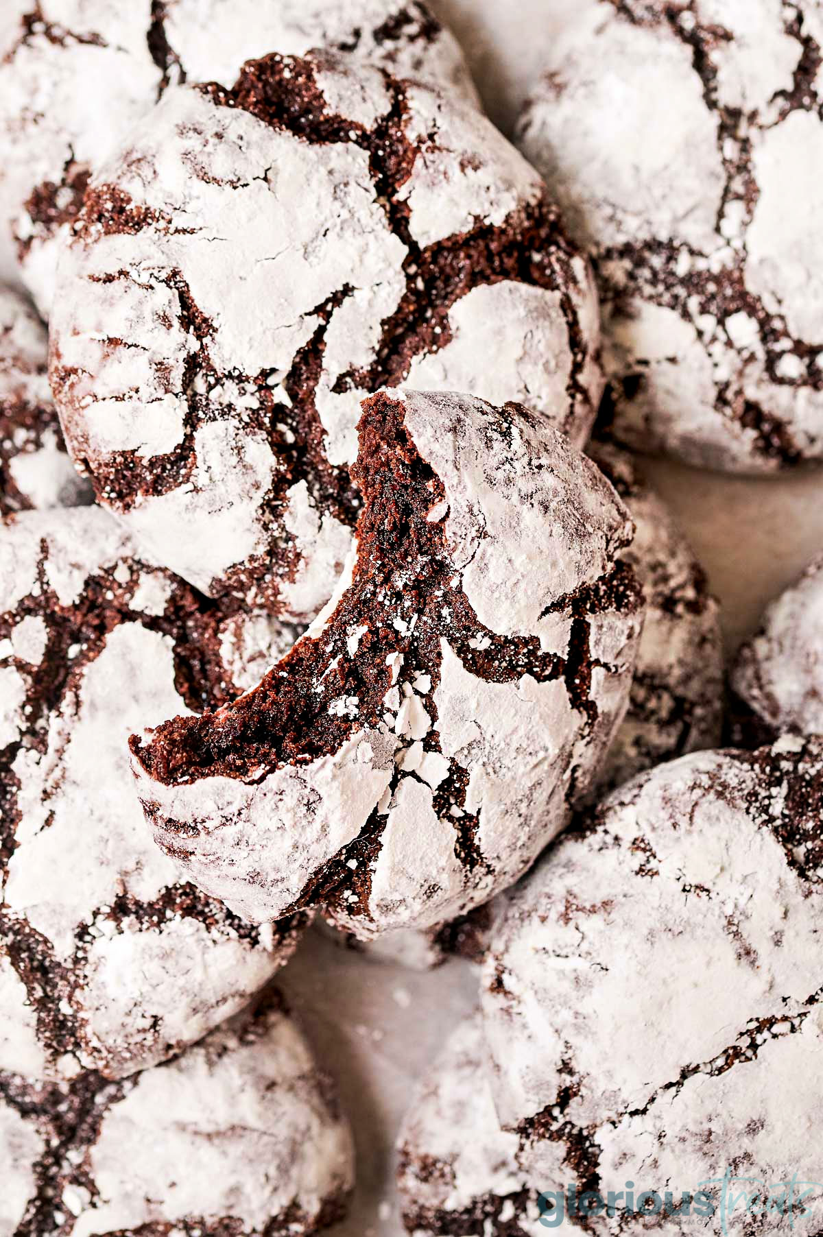 top down look at chocolate crinkle cookies with the classic powdered sugar finish. top cookie has a bite taken out of it.