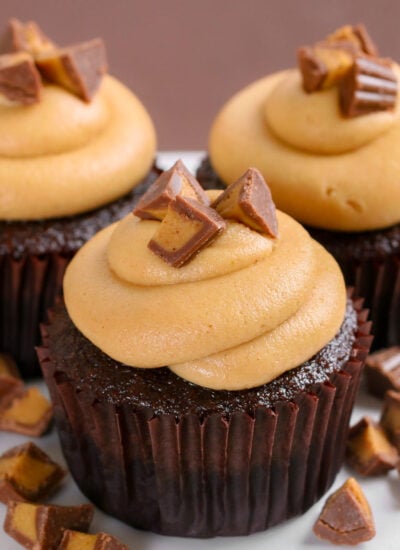 three chocolate cupcakes with swirled peanut butter frosting and chopped Reeses candy on top sitting on a white cake stand.