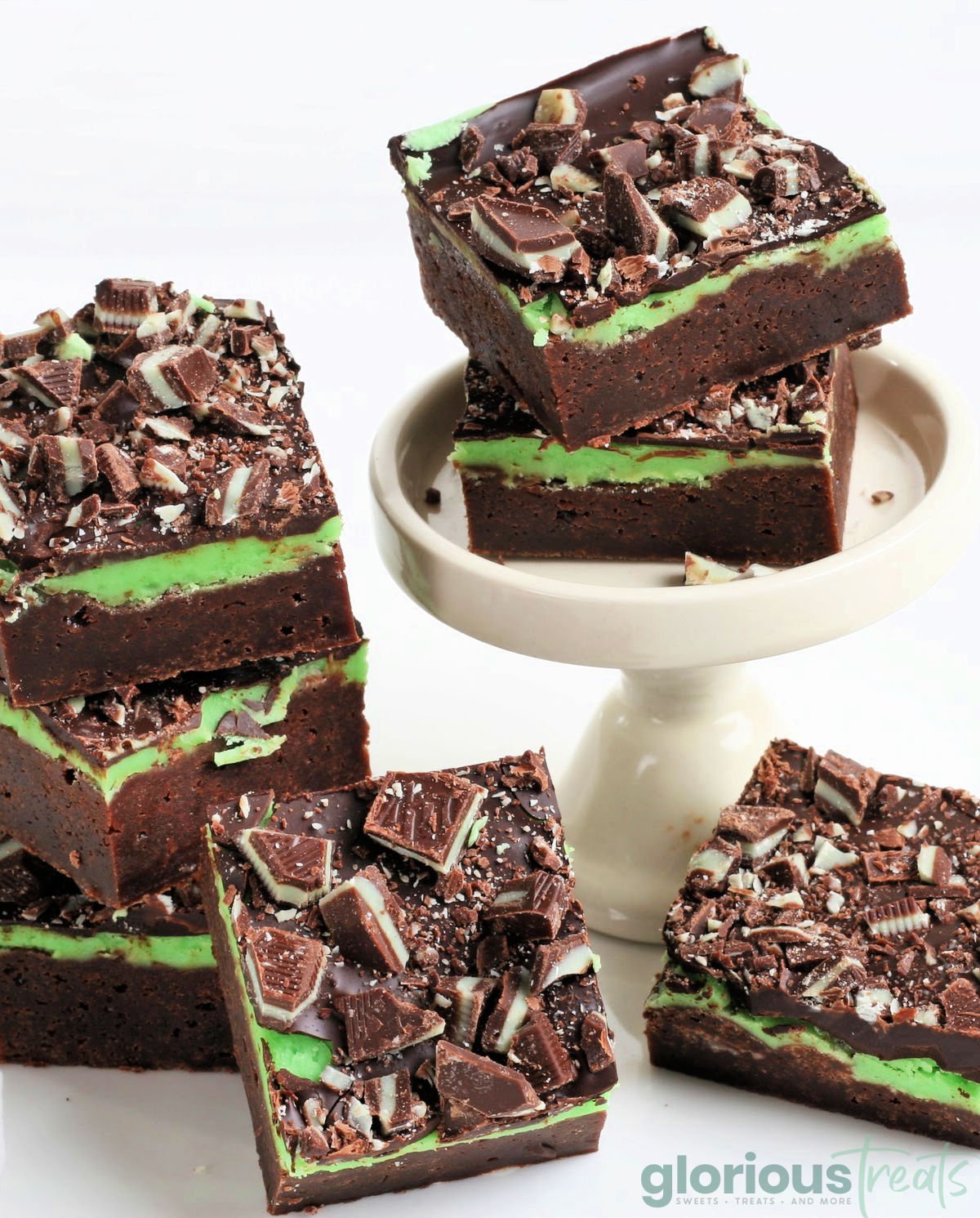 mint brownie stacked on small white pedestal and scattered around the white surface below the pedestal. 