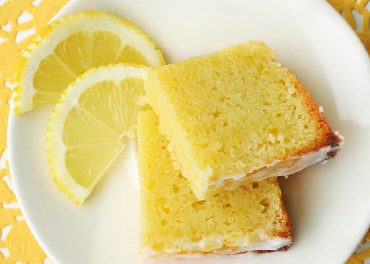 slice of lemon bread cut in half with fresh lemon slices on the side on a white plate with yellow placemat underneath