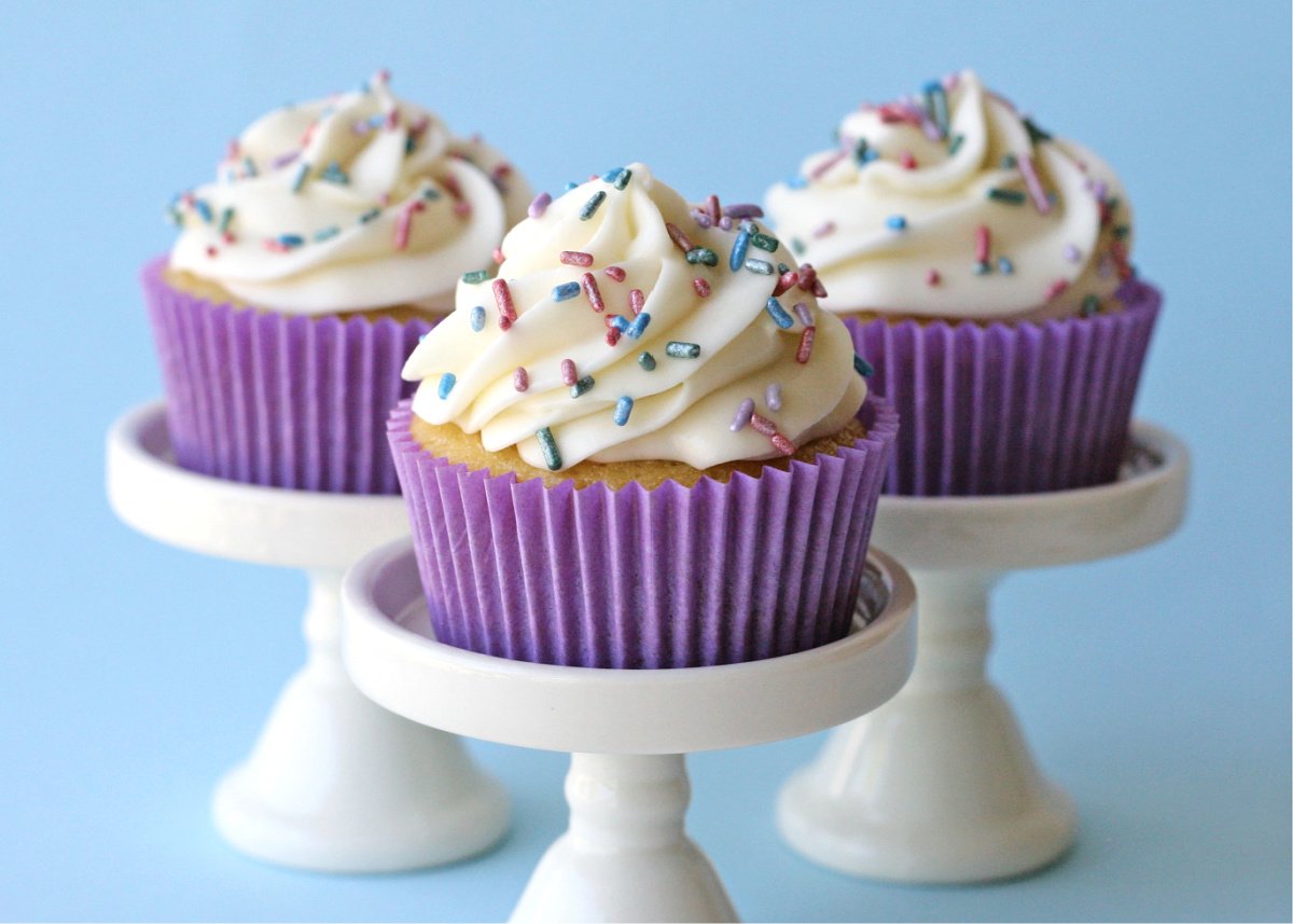 three vanilla cupcakes on white cupcake stands with purple cupcake liners and vanilla frosting