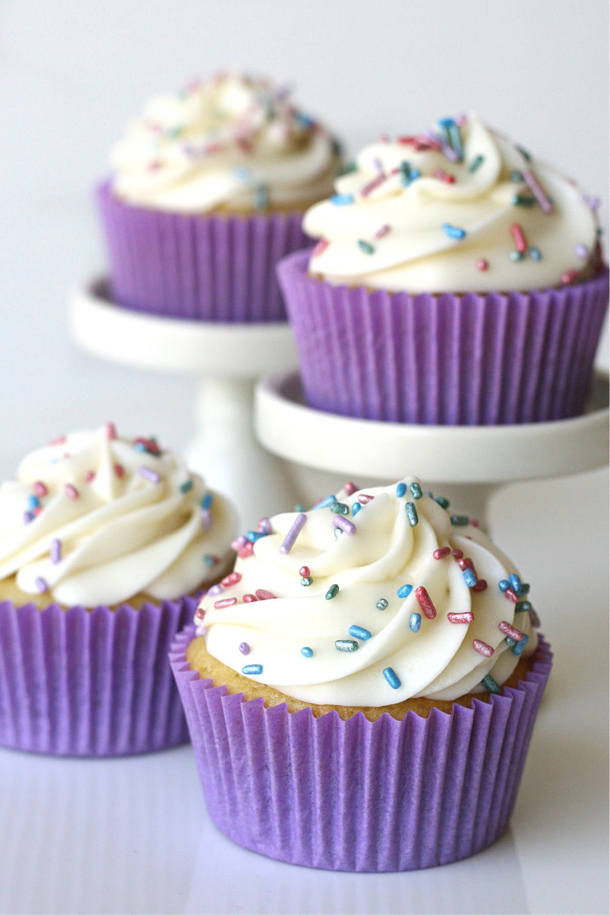 vanilla cupcakes frosted with pastel sprinkles two cupcakes sitting on white cake stands with two more cupcakes sitting below them