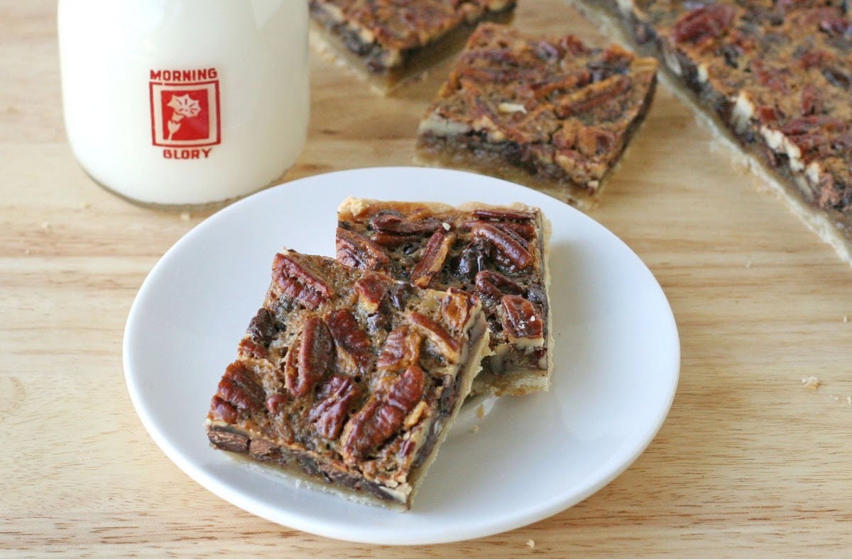 two chocolate pecan pie bars on small white plate sitting in front of rest of pan of pie bars and a glass of milk.