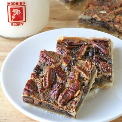 two pecan pie bars with chocolate chips sitting on small white plate in front of a glass of milk.