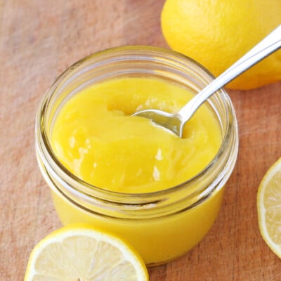 hoememade lemon curd in small glass jar with fresh lemon slices and spoon in jar square