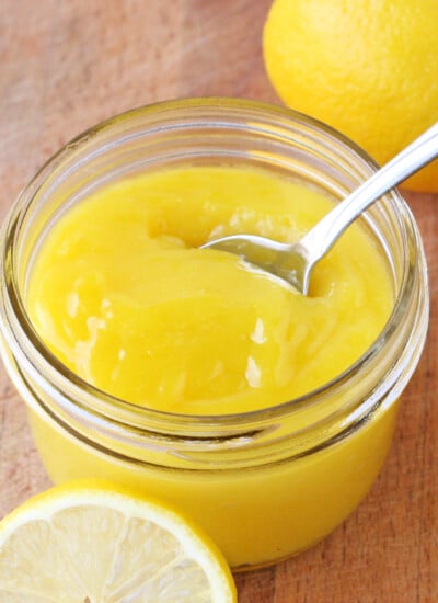 hoememade lemon curd in small glass jar with fresh lemon slices and spoon in jar square