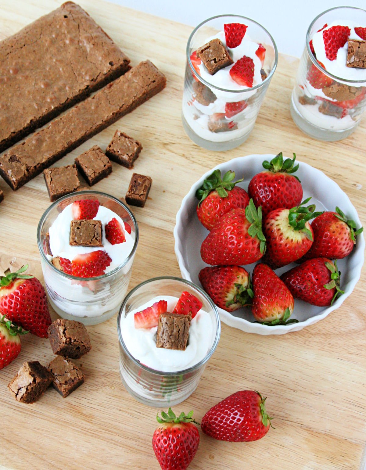 brownie strawberry shortcakes assembled in glasses with ingredients around them on a wood board.