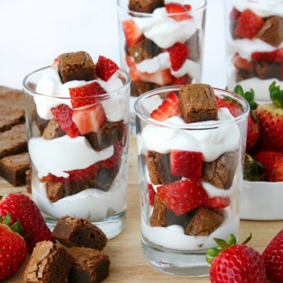 three small glasses with brownies chunks, whipped cream and strawberries layered in them.