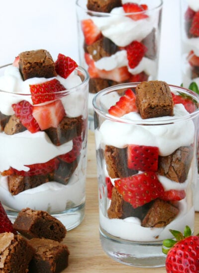 three small glasses with brownies chunks, whipped cream and strawberries layered in them.