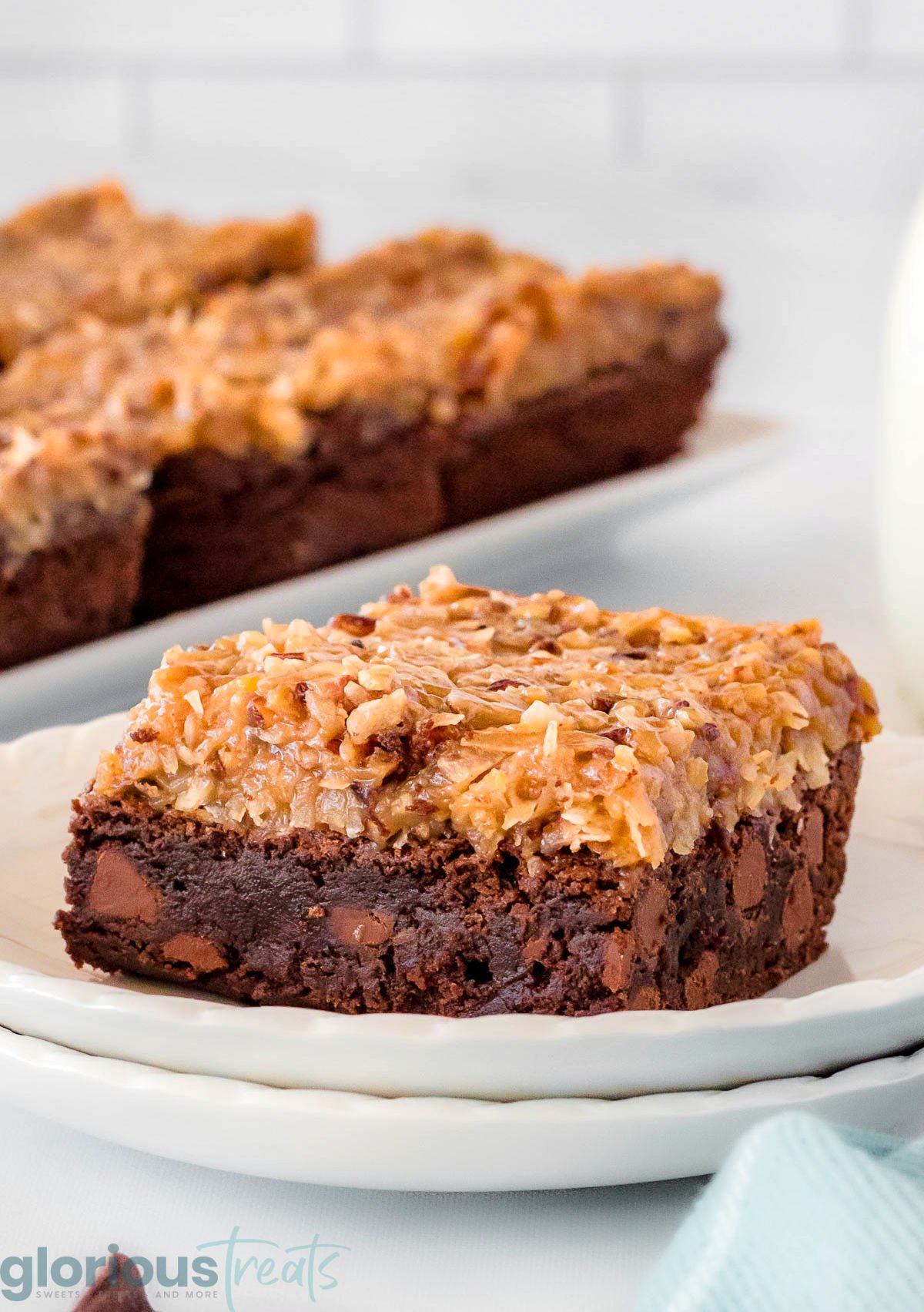german chocolate brownie sitting on white round plate with more brownies seen in background. the brownies are topped with the classic coconut pecan frosting.