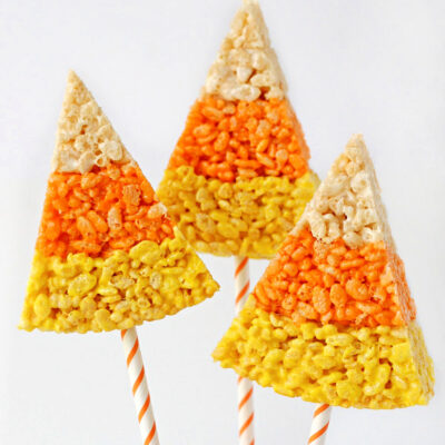 three wedge shaped rice krispie treats made to look like candy corn with straws inserted in the bottom. sitting in a white bucket filled with candy corn.