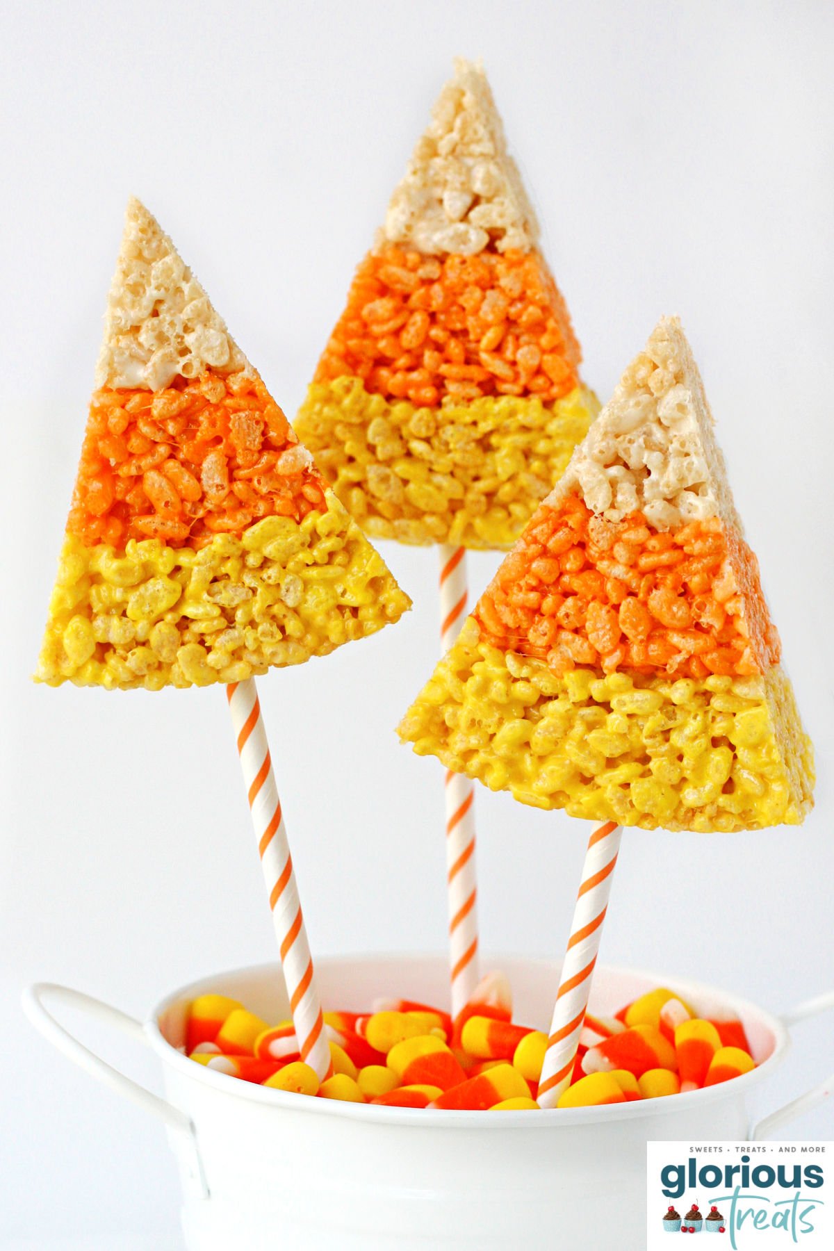 three wedge shaped rice krispie treats made to look like candy corn with straws inserted in the bottom. sitting in a white bucket filled with candy corn.