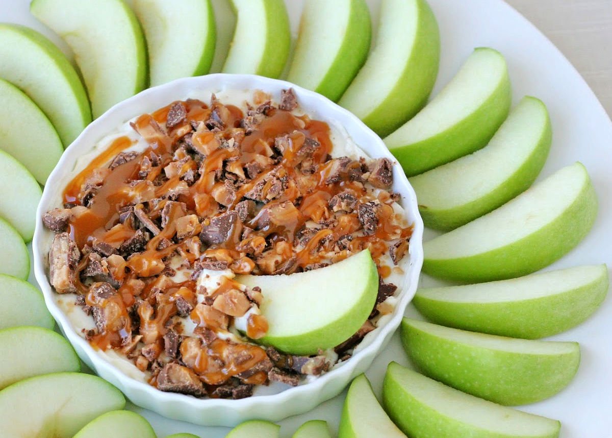 caramel apple dip in white bowl with green apple slices surrounding the bowl and one apple in the dip.