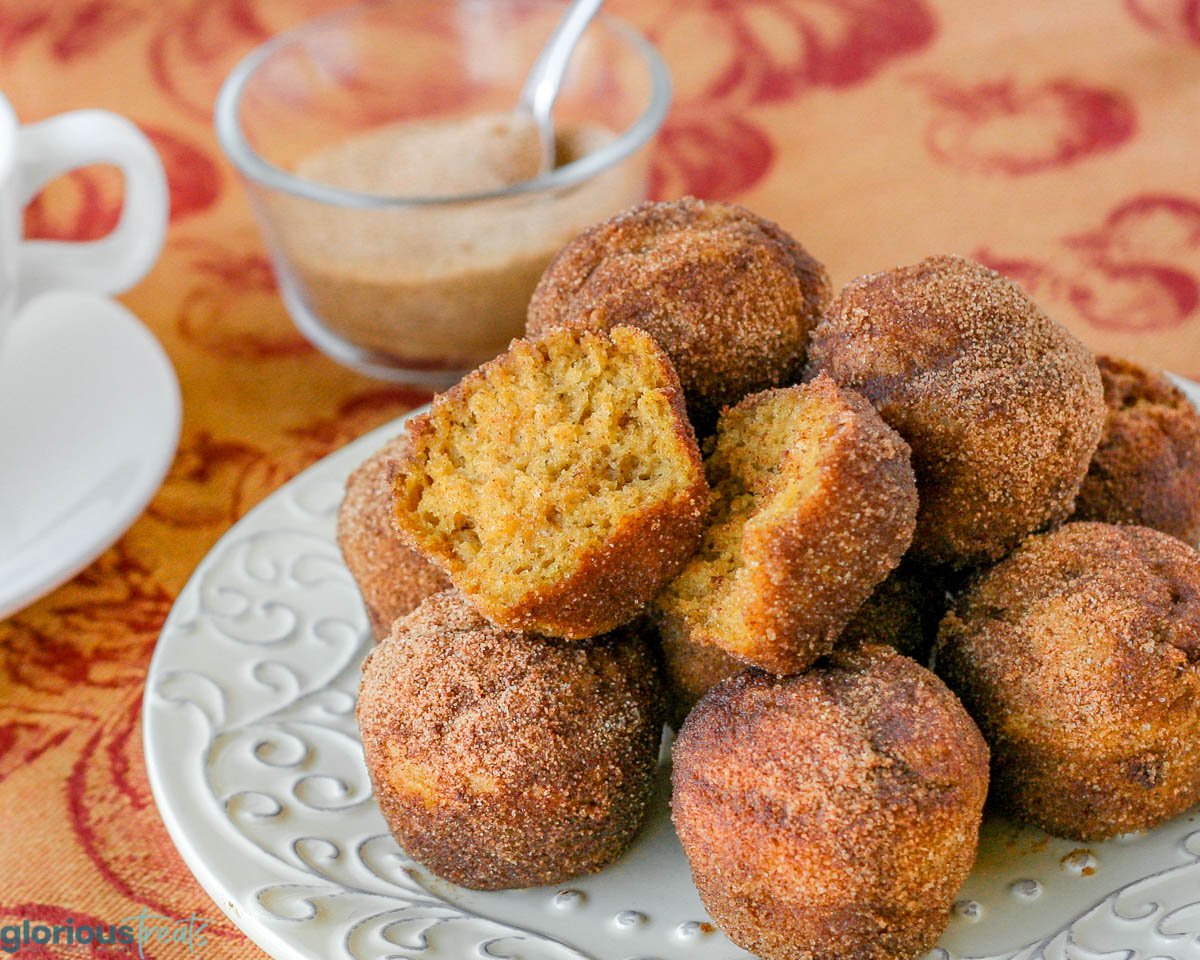 round white plate with pumpkin donut muffins piled on top. white coffee cup to the left with cinnamon sugar bowl in the background.