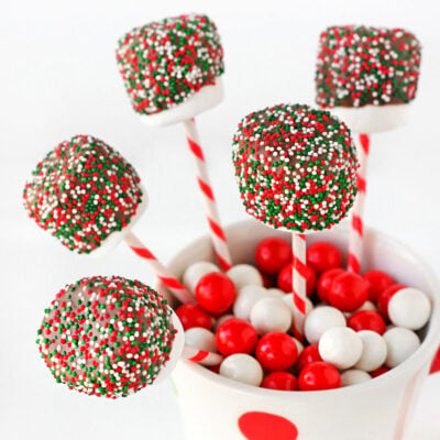 chocolate covered marshmallow pops decorated with christmas sprinkles tucked into a candy filled christmas mug.