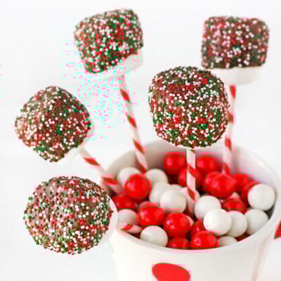 chocolate covered marshmallow pops decorated with christmas sprinkles tucked into a candy filled christmas mug.