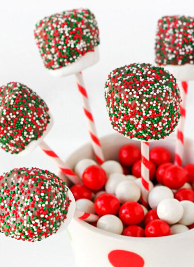 chocolate covered marshmallow pops decorated with christmas sprinkles tucked into a candy filled christmas mug.