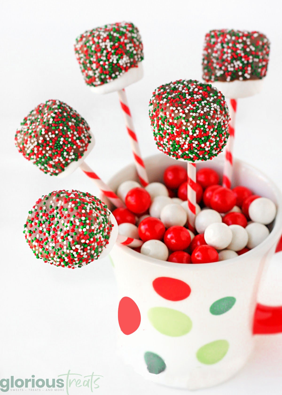 chocolate covered marshmallow pops decorated with christmas sprinkles tucked into a candy filled christmas mug.