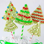 christmas rice krispie treats in the shape of trees with lollipop sticks stuck in the bottom resting in green bucket. rice krispie treats are decorated like christmas tress.