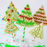 christmas rice krispie treats in the shape of trees with lollipop sticks stuck in the bottom resting in green bucket. rice krispie treats are decorated like christmas tress.