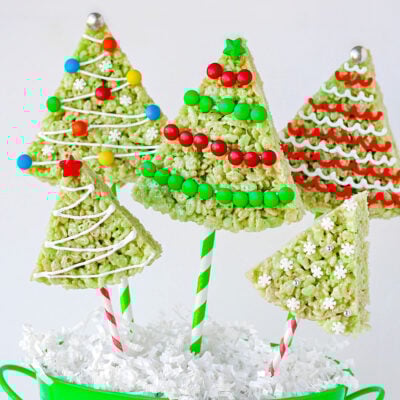 christmas rice krispie treats in the shape of trees with lollipop sticks stuck in the bottom resting in green bucket. rice krispie treats are decorated like christmas tress.