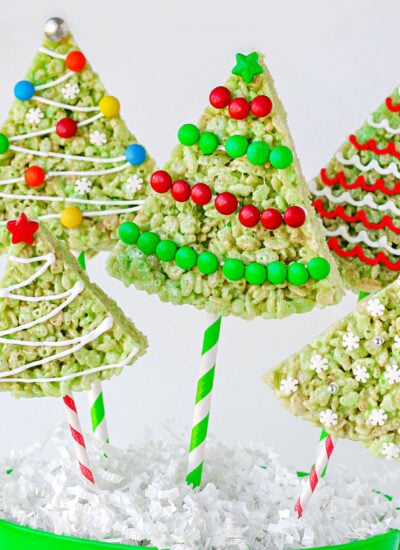 christmas rice krispie treats in the shape of trees with lollipop sticks stuck in the bottom resting in green bucket. rice krispie treats are decorated like christmas tress.