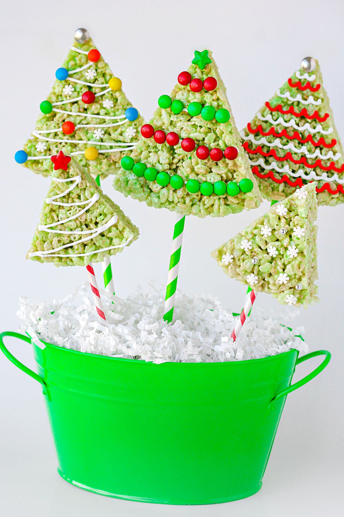 christmas rice krispie treats in the shape of trees with lollipop sticks stuck in the bottom resting in green bucket. rice krispie treats are decorated like christmas tress.