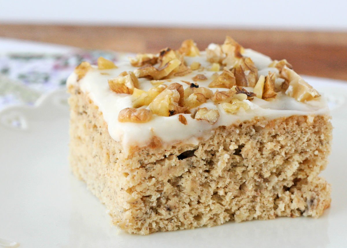 front view of a piece of banana cake on white plate topped with frosting and nuts.