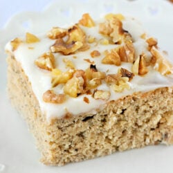 piece of banana cake with cream cheese frosting and walnuts on top sitting on white plate.
