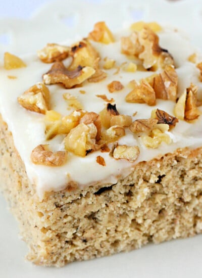 piece of banana cake with cream cheese frosting and walnuts on top sitting on white plate.