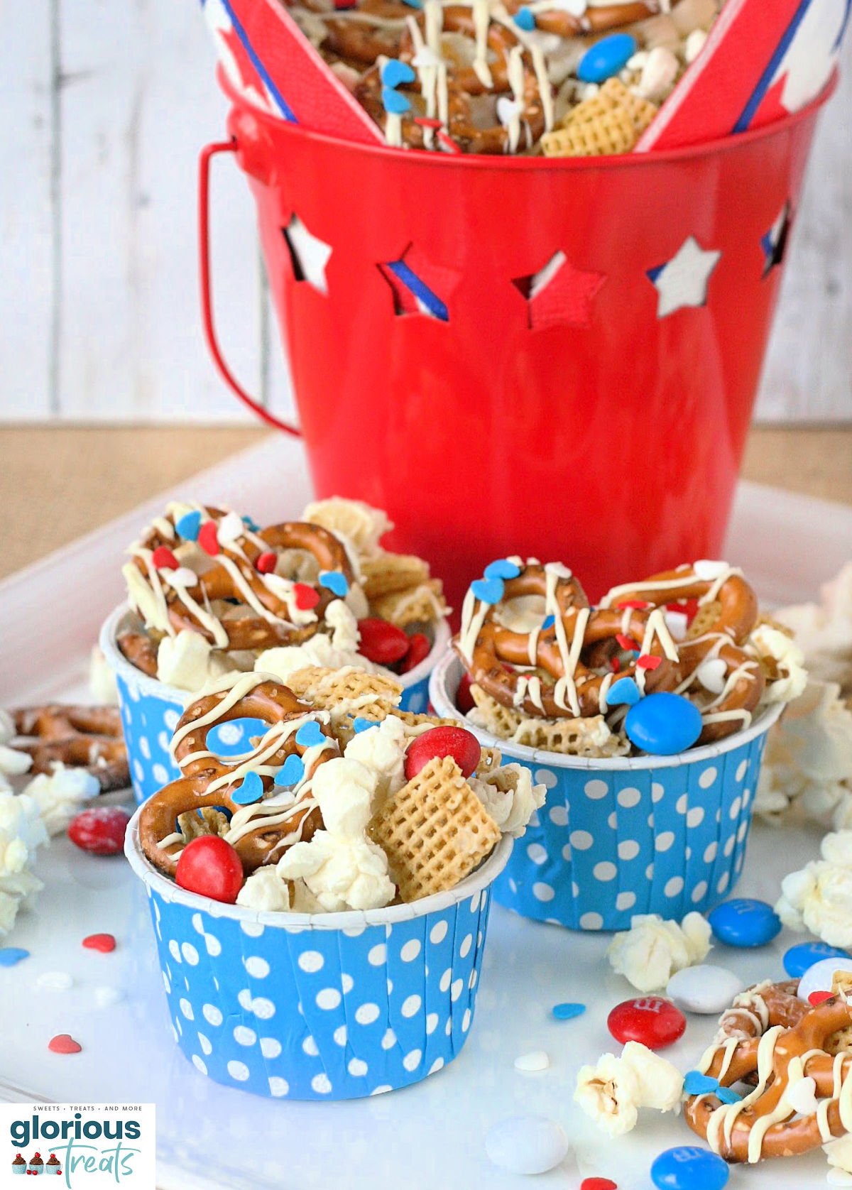 4th of July snack mix made with popcorn, pretzels and candy in small blue and white polka dot paper cups sitting on white plate.
