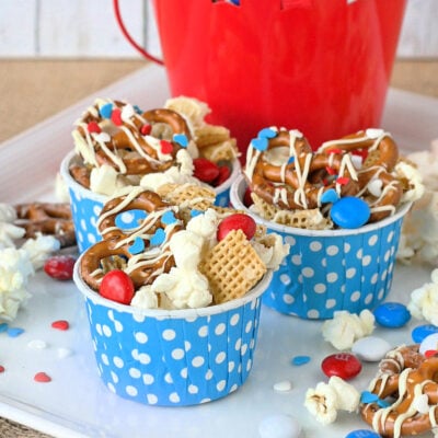 4th of july snack mix made with popcorn, pretzels and candy in small blue and white polka dot paper cups sitting on white plate.