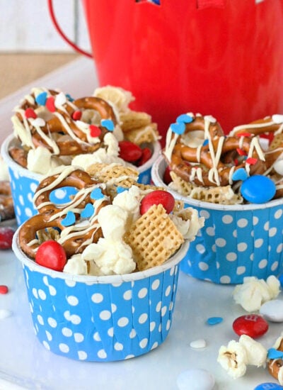 4th of july snack mix made with popcorn, pretzels and candy in small blue and white polka dot paper cups sitting on white plate.