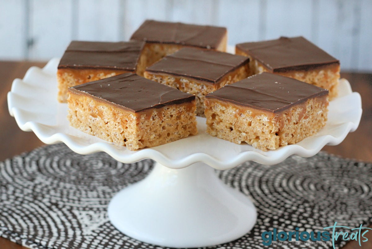 six rice krispie treats made with caramel and topped with chocolate are on a white cake stand with a fluted edge.