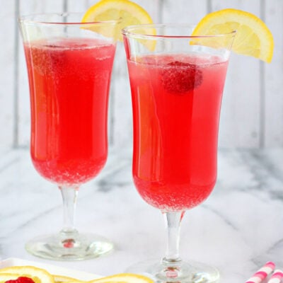 two punch glasses filled with bright pink party punch and topped with a lemon slice and fresh raspberry.