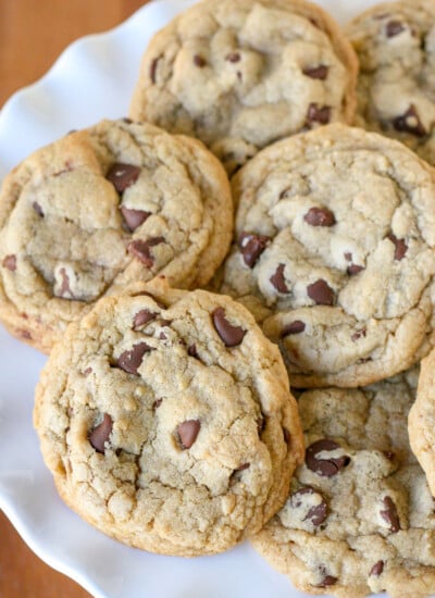 chocolate chip cookies piled on white cake stand with wood surface underneath square image