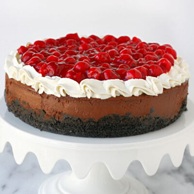 whole black forest cheesecake topped with whipped cream and cherry pie filling on a white metal cake stand.