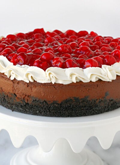 whole black forest cheesecake topped with whipped cream and cherry pie filling on a white metal cake stand.