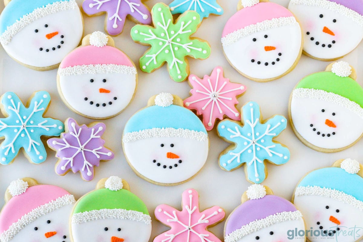 Sugar cookies cut out into snowman faces and decorated with royal icing. The snowman caps are pink, blue and purple. Snowflake cookies in the same colors are decorated and also are on the tray.