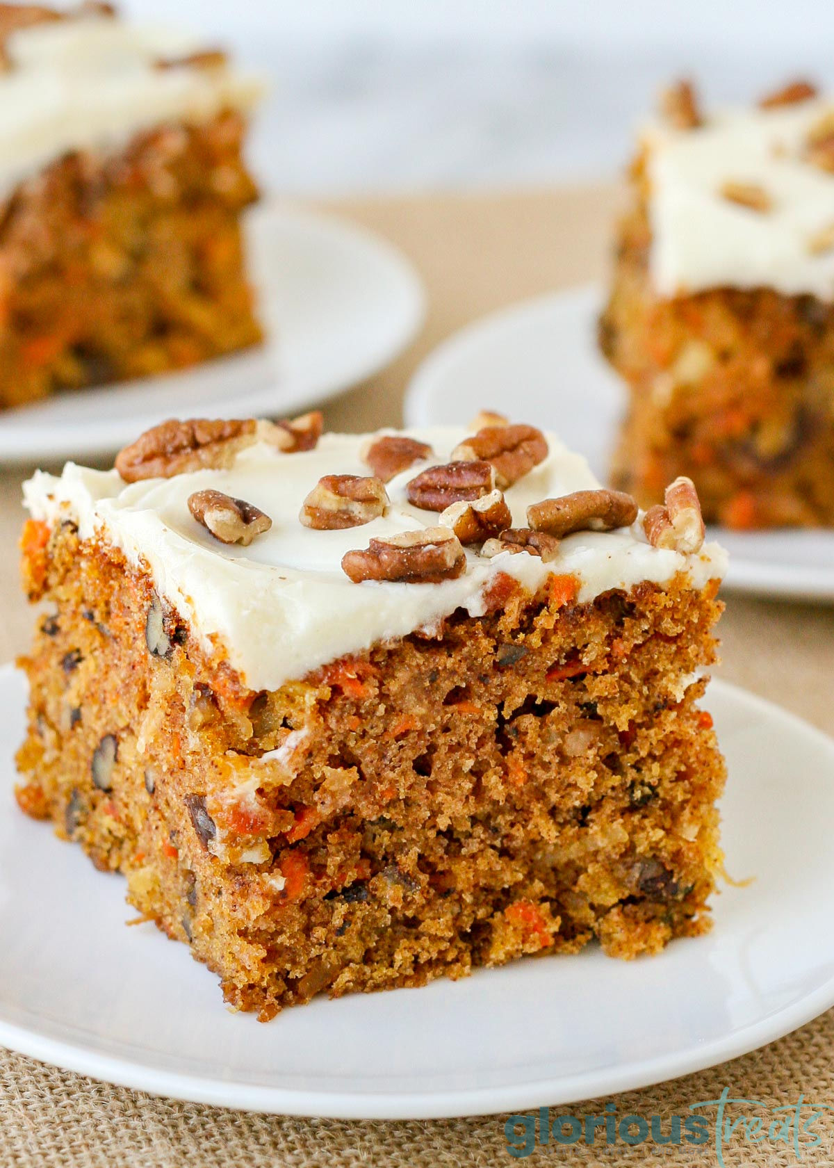 one square piece of carrot cake topped with cream cheese frosting and chopped pecans. cake is on a small white plate with two more plates in the background.