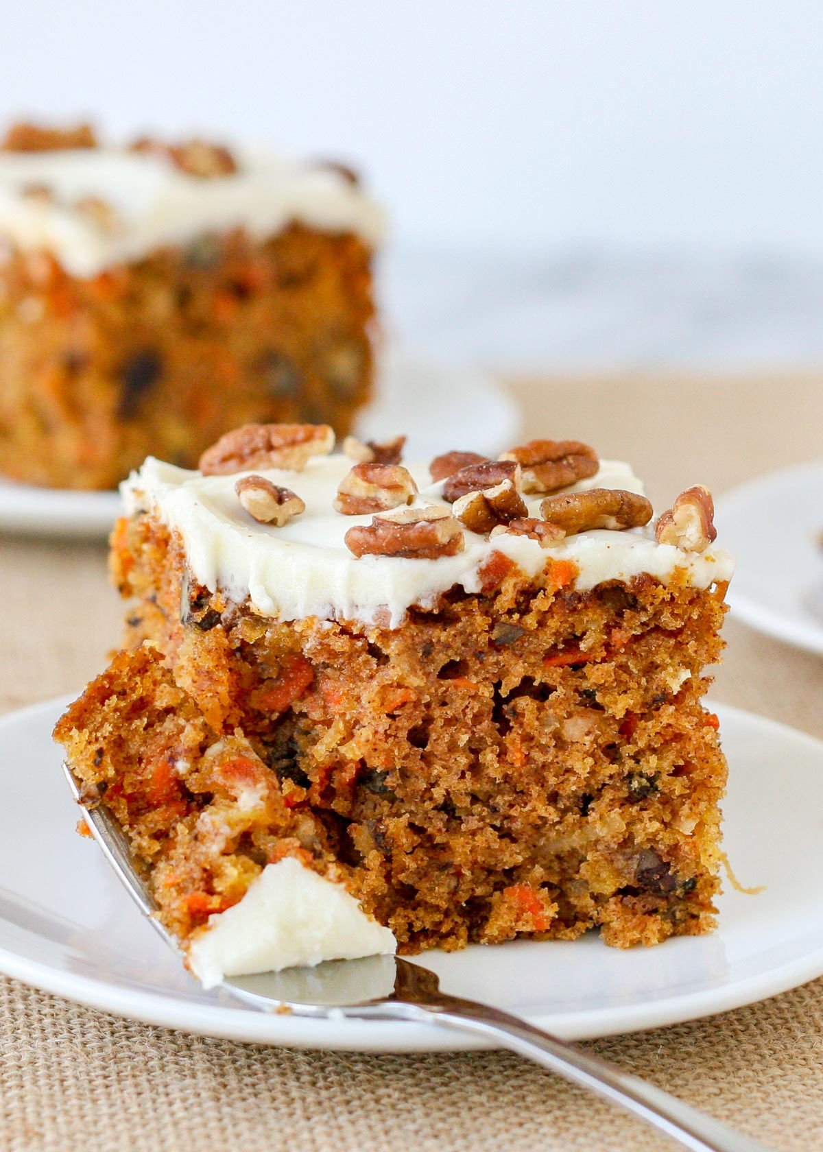 piece of carrot cake with a bite taken and the fork resting on the side of the white plate. two more pieces of carrot cake can be seen in the background.