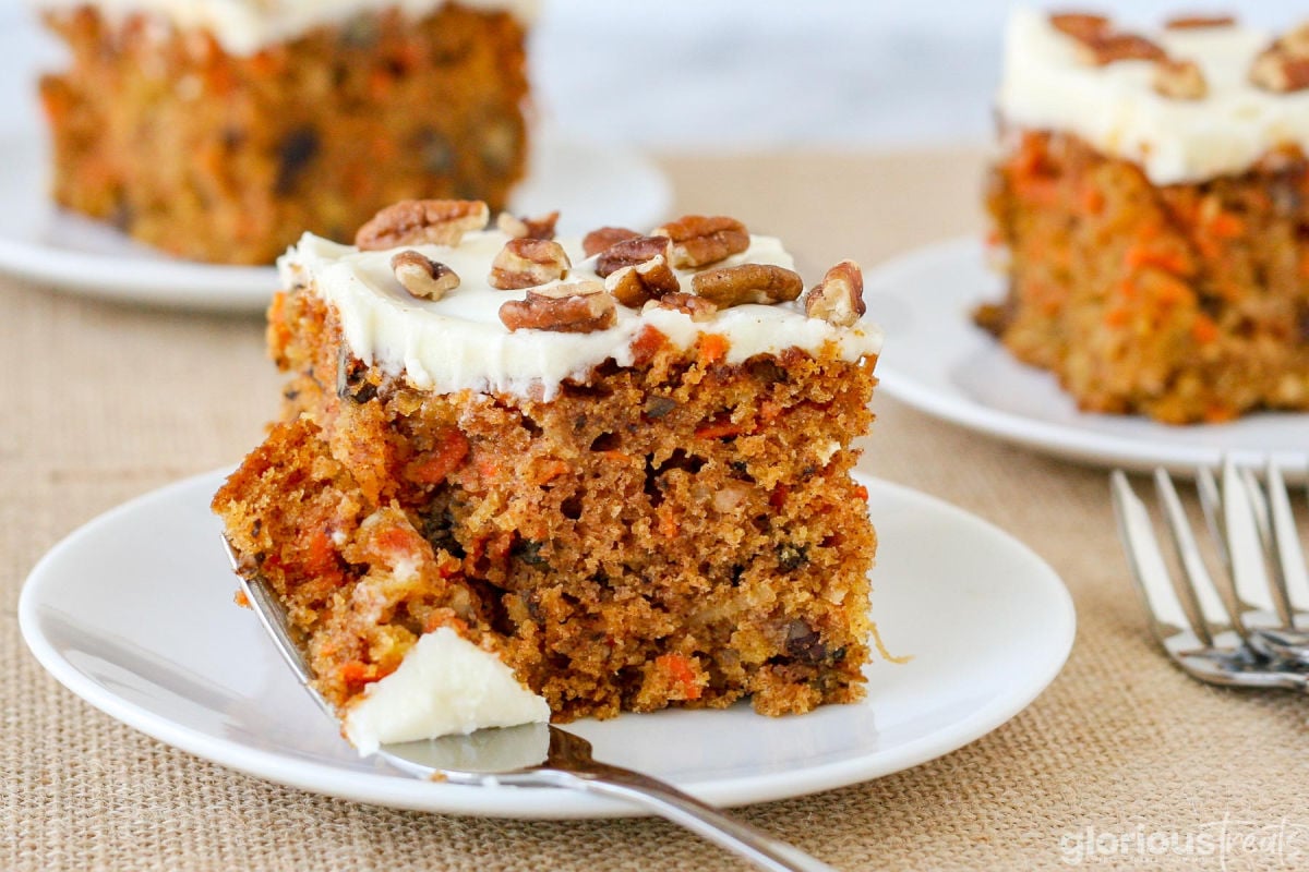 piece of carrot cake with a bite taken and the fork resting on the side of the white plate. two more pieces of carrot cake can be seen in the background.