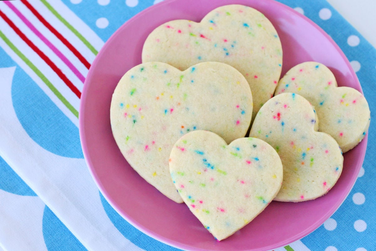 sugar cookies with sprinkles cut into heart shapes on pink plate with blue napkin beneath