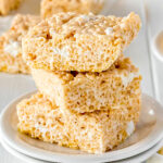 three rice krispie treat squares stacked on a white plate with more treats in the background and marshmallows scattered around the front.