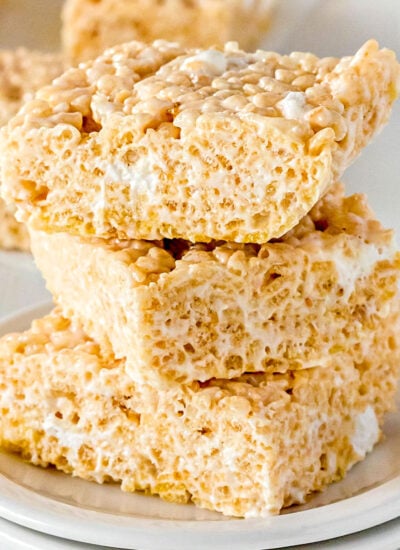 three rice krispie treat squares stacked on a white plate with more treats in the background and marshmallows scattered around the front.