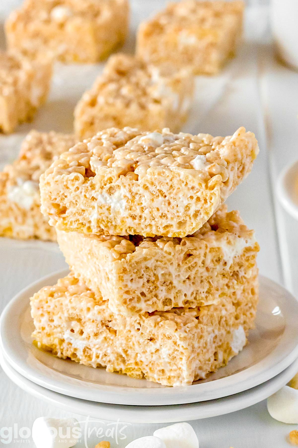 three rice krispie treat squares stacked on a white plate with more treats in the background and marshmallows scattered around the front. 