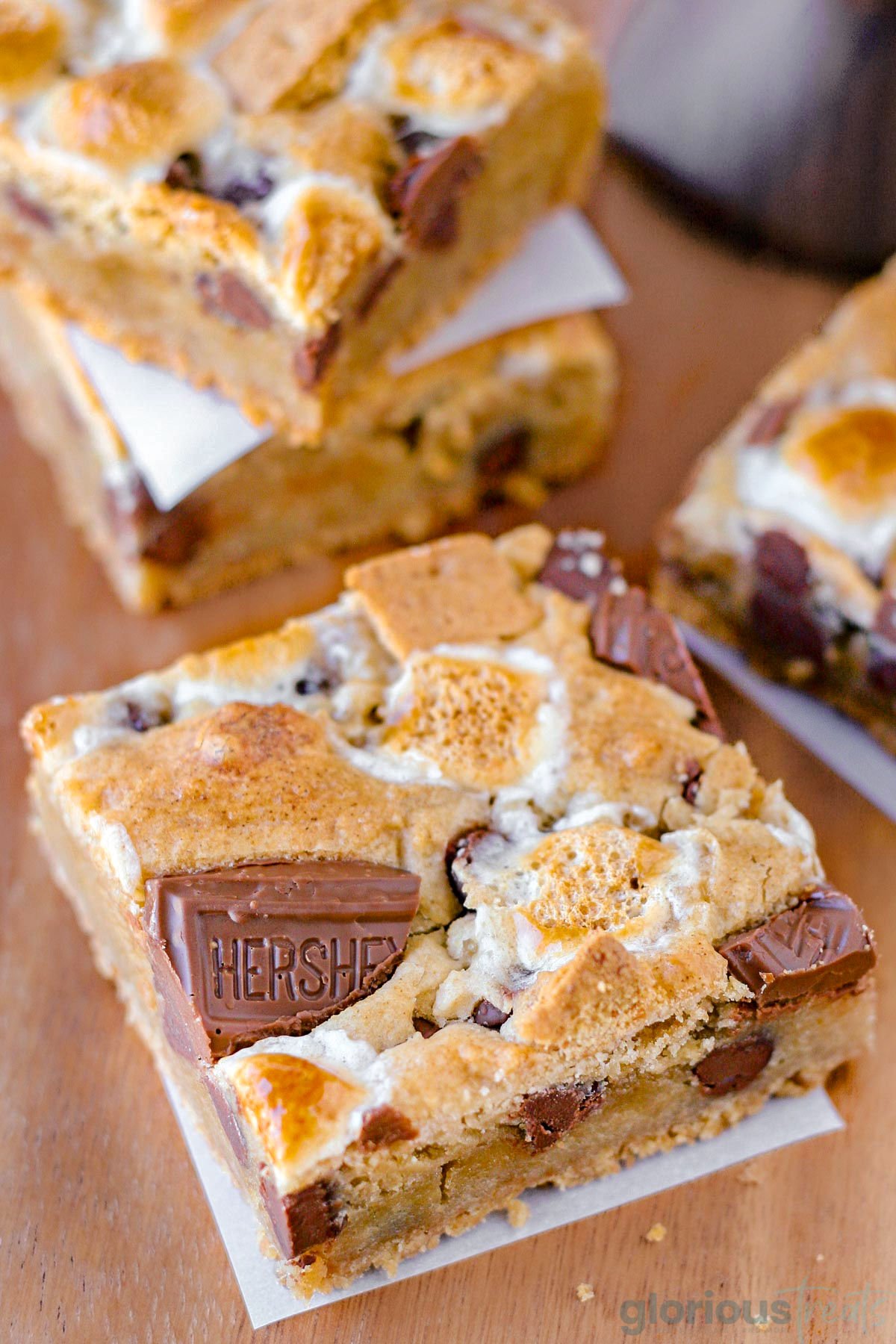Smores bar recipe showing cut into squares and layered with parchment paper. Single bar sitting up front on wood surface.