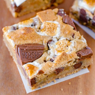 Smores bar recipe showing cut into squares and layered with parchment paper. Single bar sitting up front on wood surface.