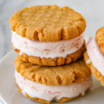 two peanut butter cookie ice cream sandwiches stacked when another sandwich leaning against them on a white plate.